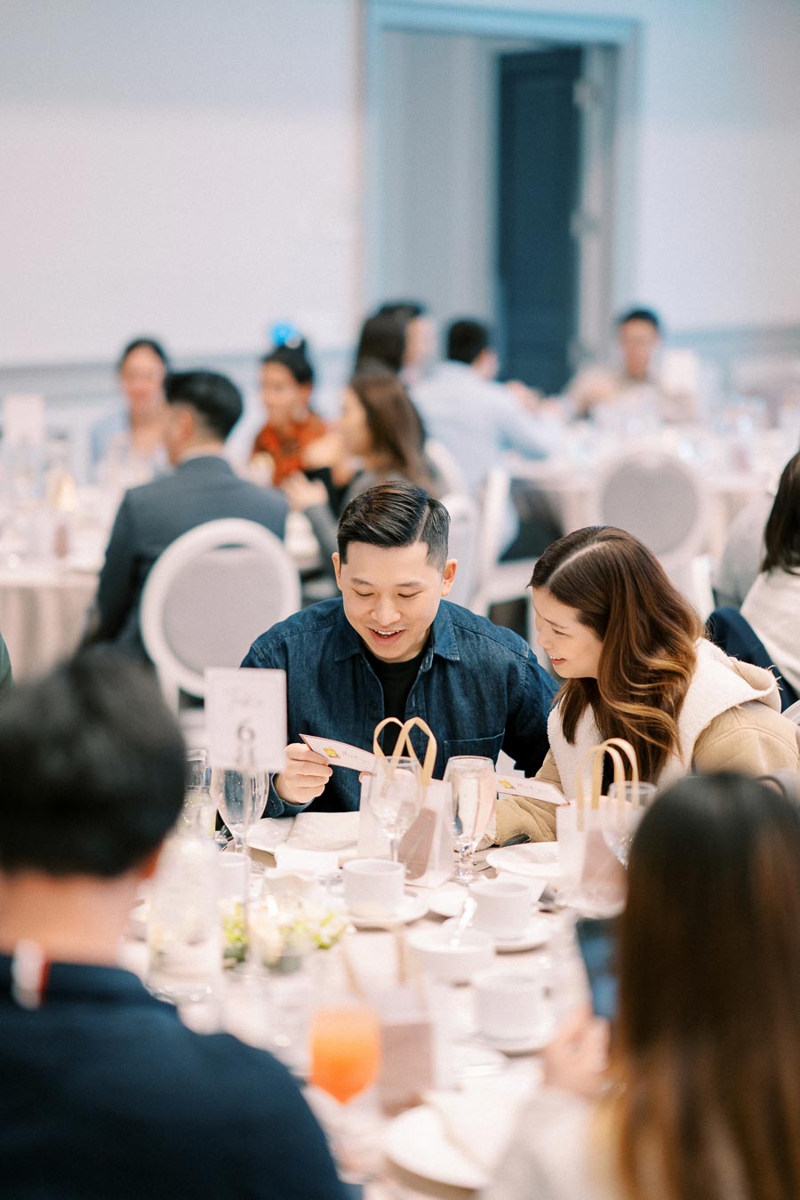 A group of people sitting around a table at a formal event, enjoying their meal and conversation. The scene captures an elegant atmosphere with tables set with white tablecloths, glasses, and decorations, indicating a special occasion or gathering.