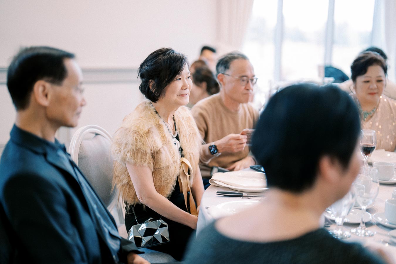 A group of elegantly dressed people seated at a dining table during a formal event, with focus on a woman wearing a fur shawl and holding a geometric purse.