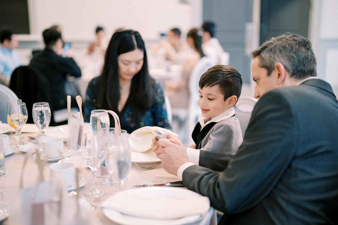 A family enjoying a meal together at a formal event, featuring a young boy in a suit interacting with an adult, surrounded by elegantly set tables and other guests in the background.