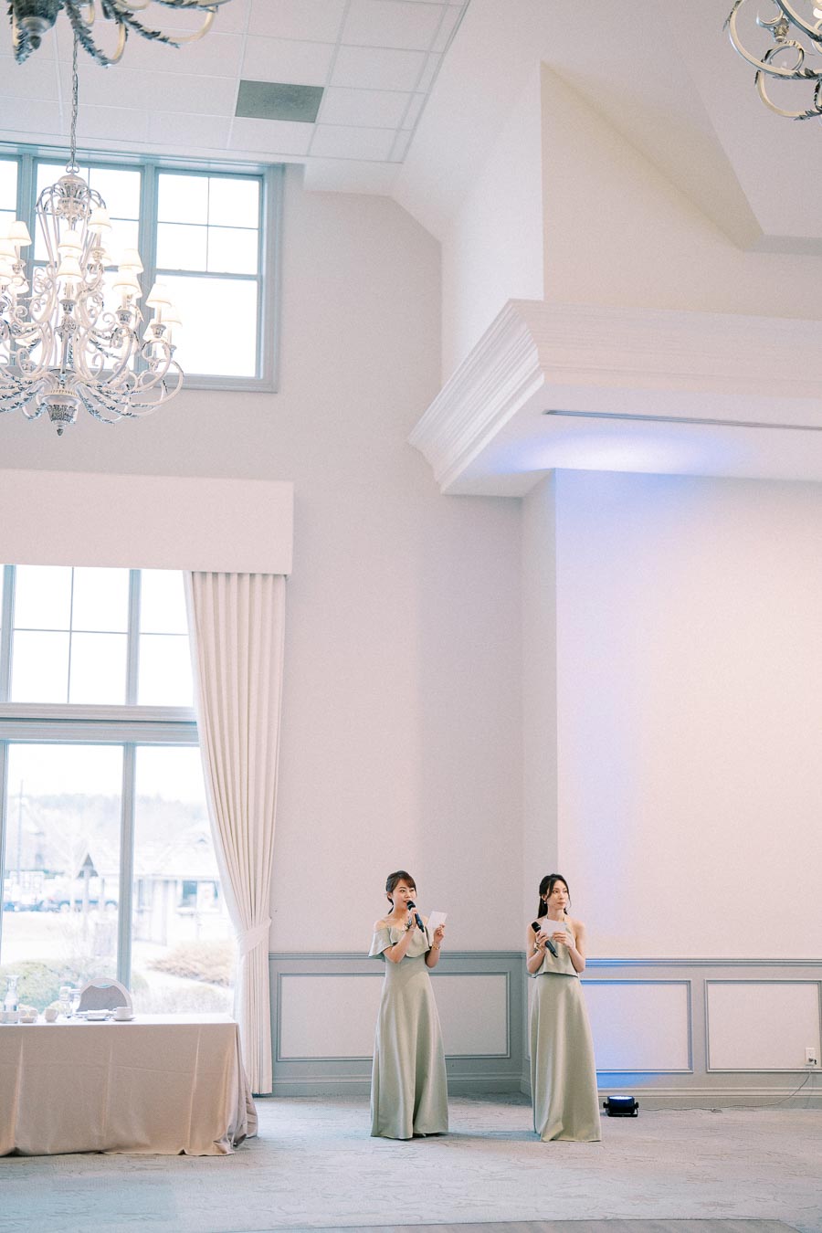 Two women in elegant, light-colored gowns holding microphones stand in a spacious, well-lit room with high ceilings, decorative chandeliers, and large windows with flowing curtains, suggesting a formal event or wedding speech setting.