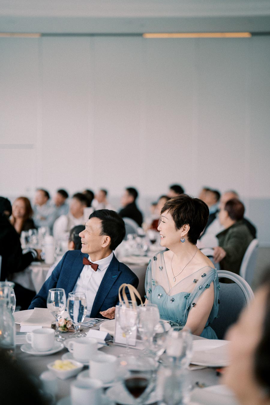 A well-dressed couple in formal attire sitting at a table, attentively listening during an elegant banquet or wedding reception. The room is filled with other attendees, elegantly set tables, and soft lighting.