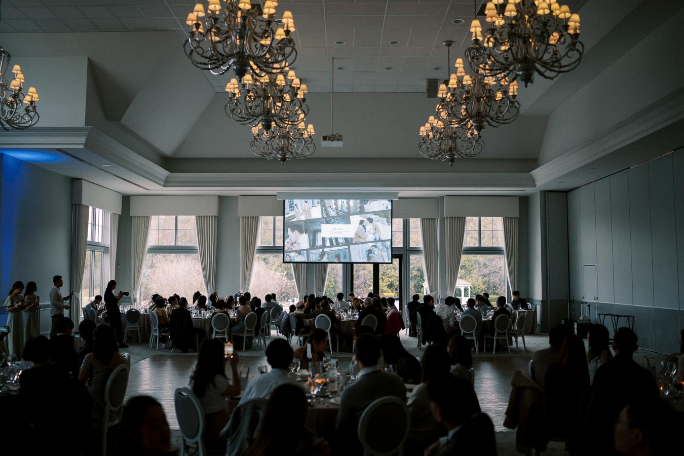 Elegant wedding reception in a large banquet hall with chandeliers, guests seated at round tables, and a large screen displaying images.