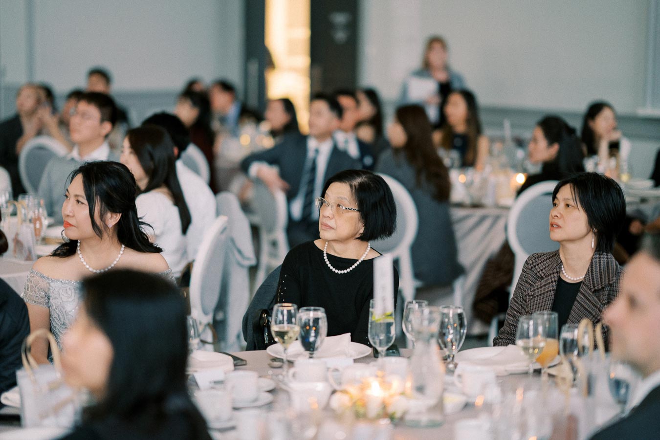 A group of elegantly dressed individuals attentively listening during a formal event or conference, seated at tables adorned with glassware and soft lighting in a banquet hall.