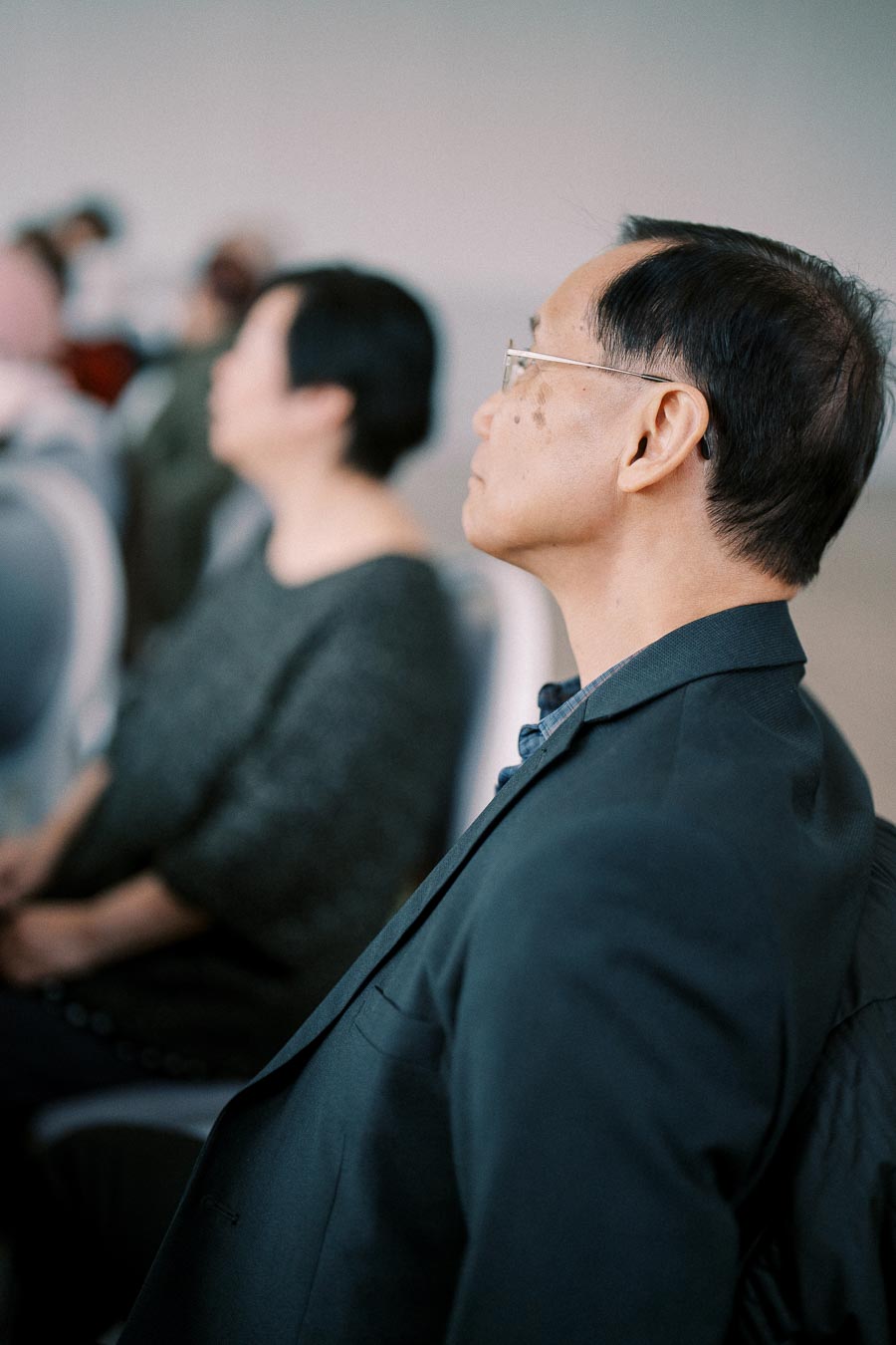 A professional man attentively listening in a business seminar, wearing glasses and a suit, with blurred audience members in the background.
