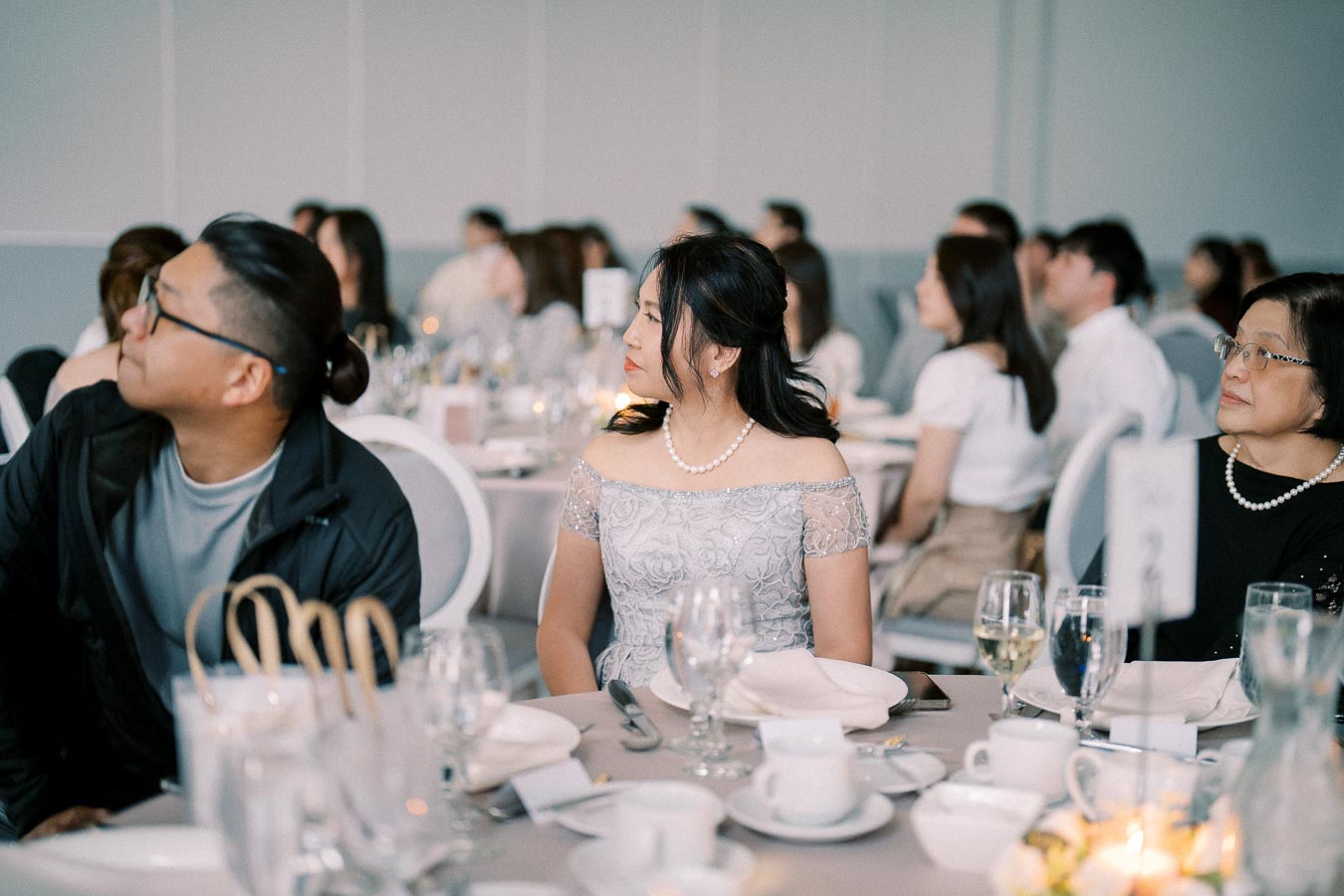 A group of people attentively listening at a formal event, seated around a table with elegant table settings, including wine glasses, plates, and silverware.