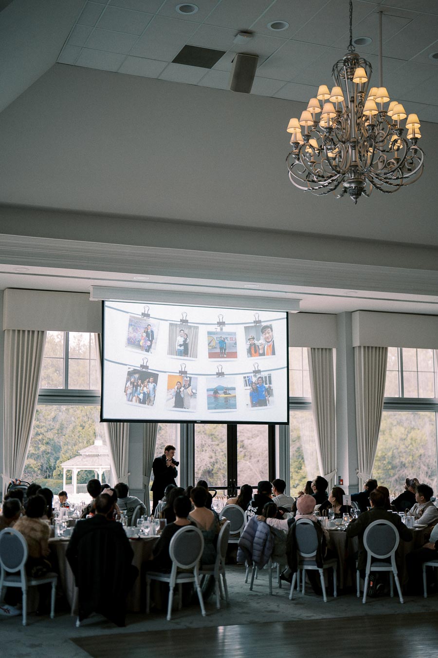 Elegant banquet hall with guests seated at round tables, viewing a large screen displaying a photo presentation, under a decorative chandelier.