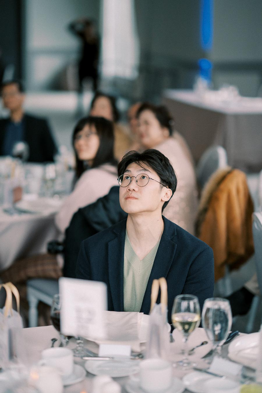 Man with glasses attentively listening during a formal event, seated among elegantly dressed guests at round tables with white table settings.