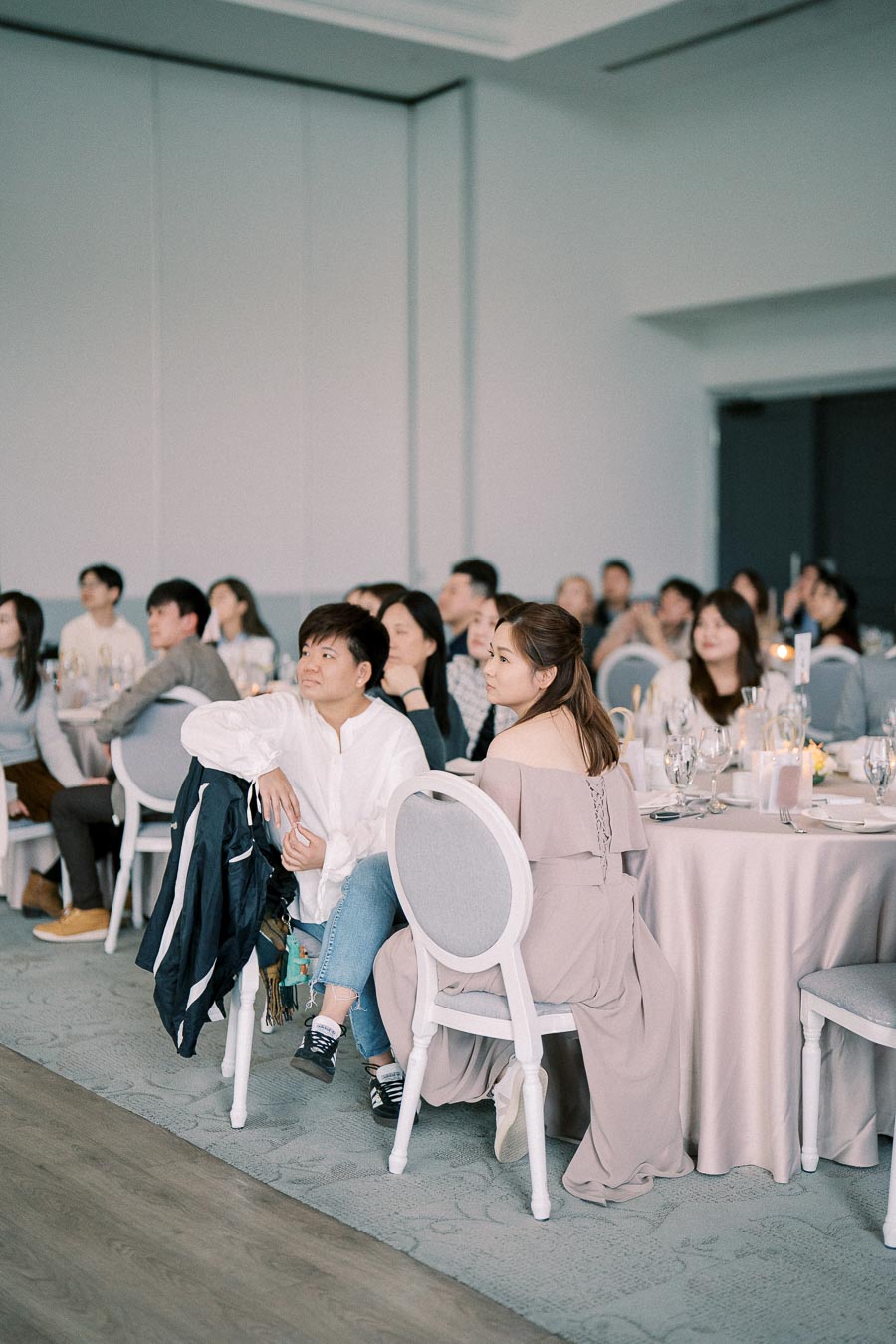 A group of people attentively watching an event in a modern, spacious venue. Tables are set with elegant tableware and floral centerpieces, creating a formal atmosphere.