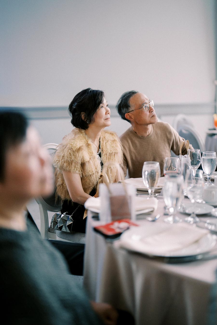 Elegantly dressed people seated at a round table attentively watching a presentation in a well-lit banquet hall.