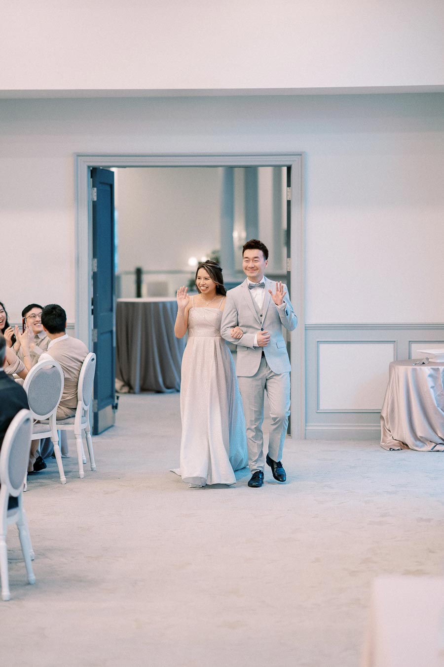 Elegant couple in formal attire making a joyful entrance at a wedding reception, smiling and waving to guests in a beautifully decorated venue.