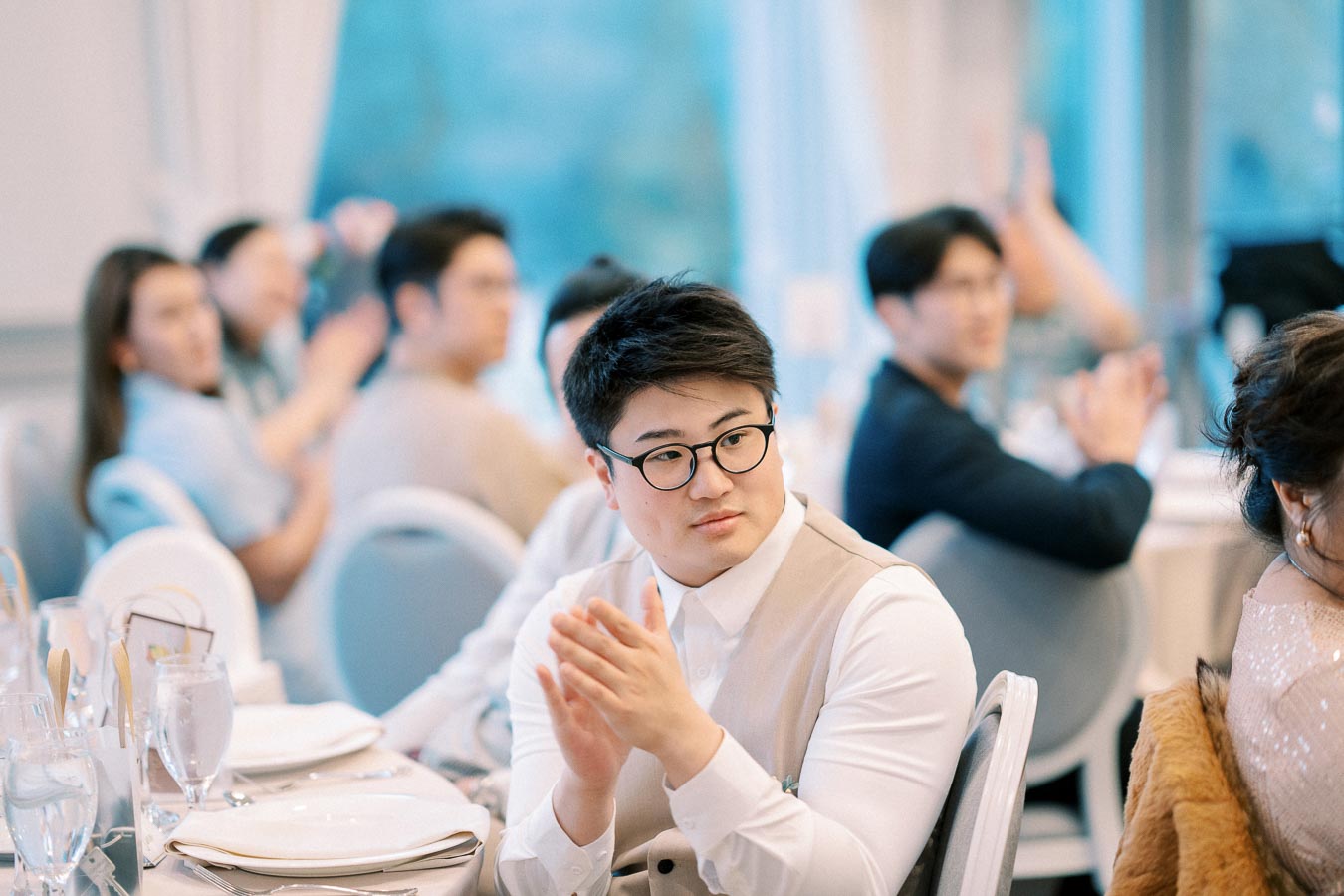 A man with glasses sitting at a formal event table, clapping, with attendees in the background.