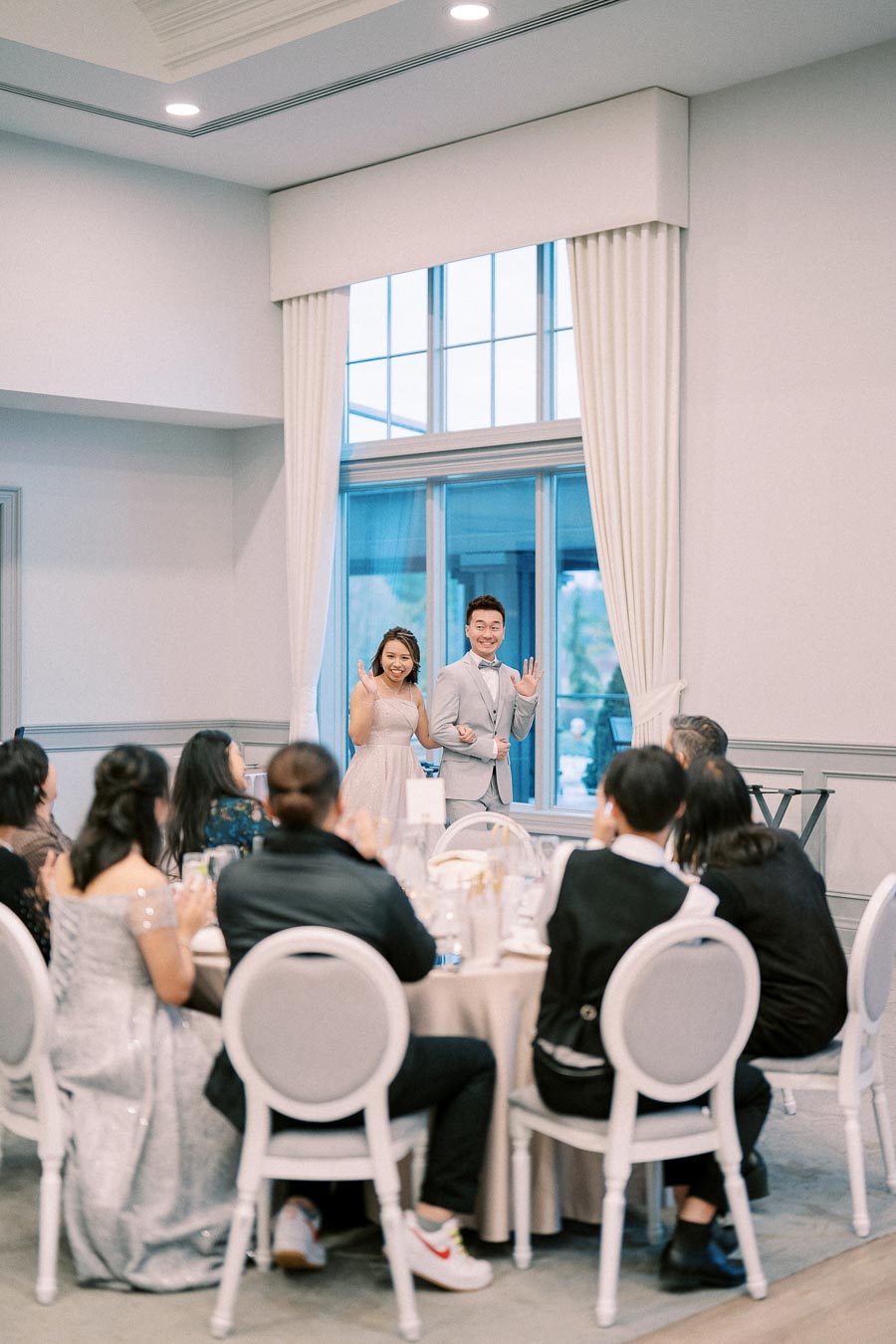 A couple dressed elegantly, smiling and waving at guests during a formal celebration in a bright, spacious venue.