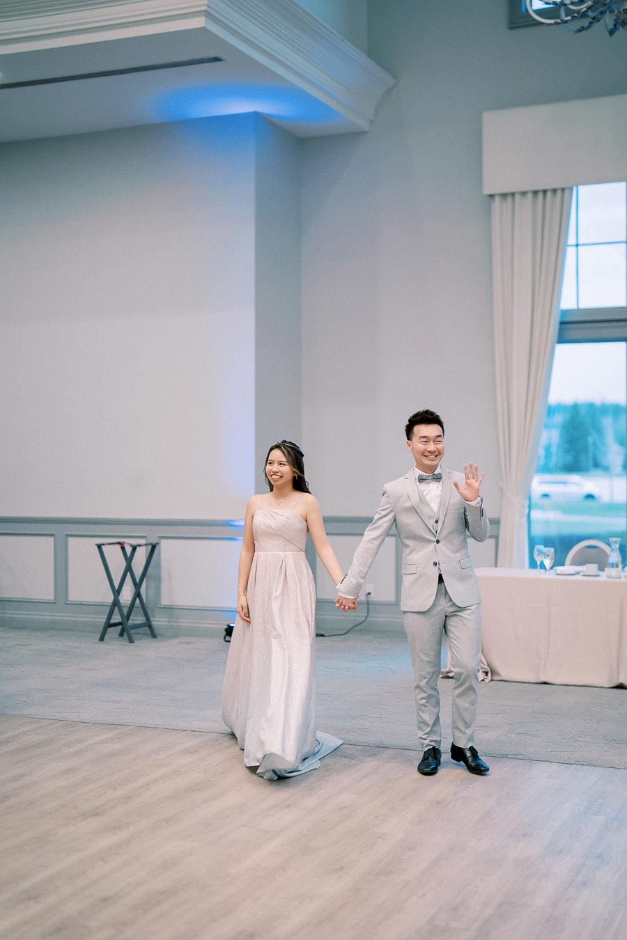 Elegant couple entering a ballroom, woman in strapless gown and man in light grey suit waving, showcasing a joyful celebration atmosphere at a formal event.