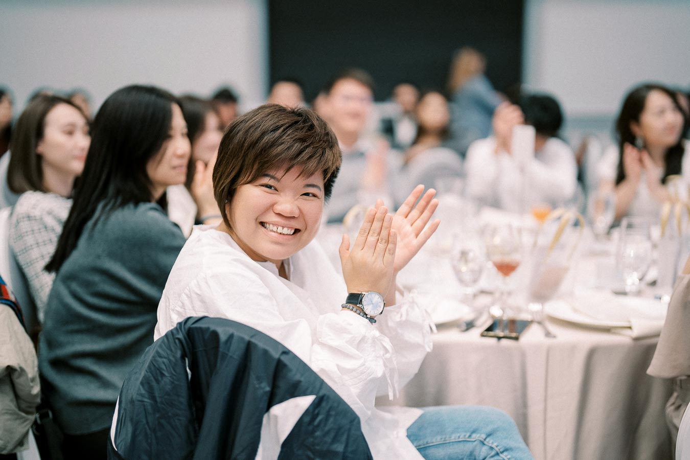 Cheerful woman clapping at a formal event with other attendees seated at a table in a conference setting.
