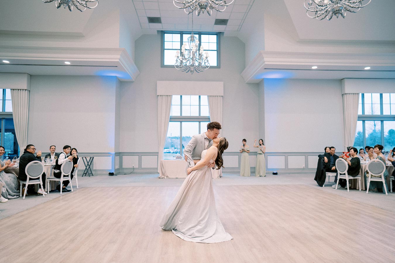 A couple shares a romantic dance in an elegant ballroom, surrounded by seated guests who are watching and clapping. Chandelier lighting enhances the modern, airy ambiance of the reception venue.