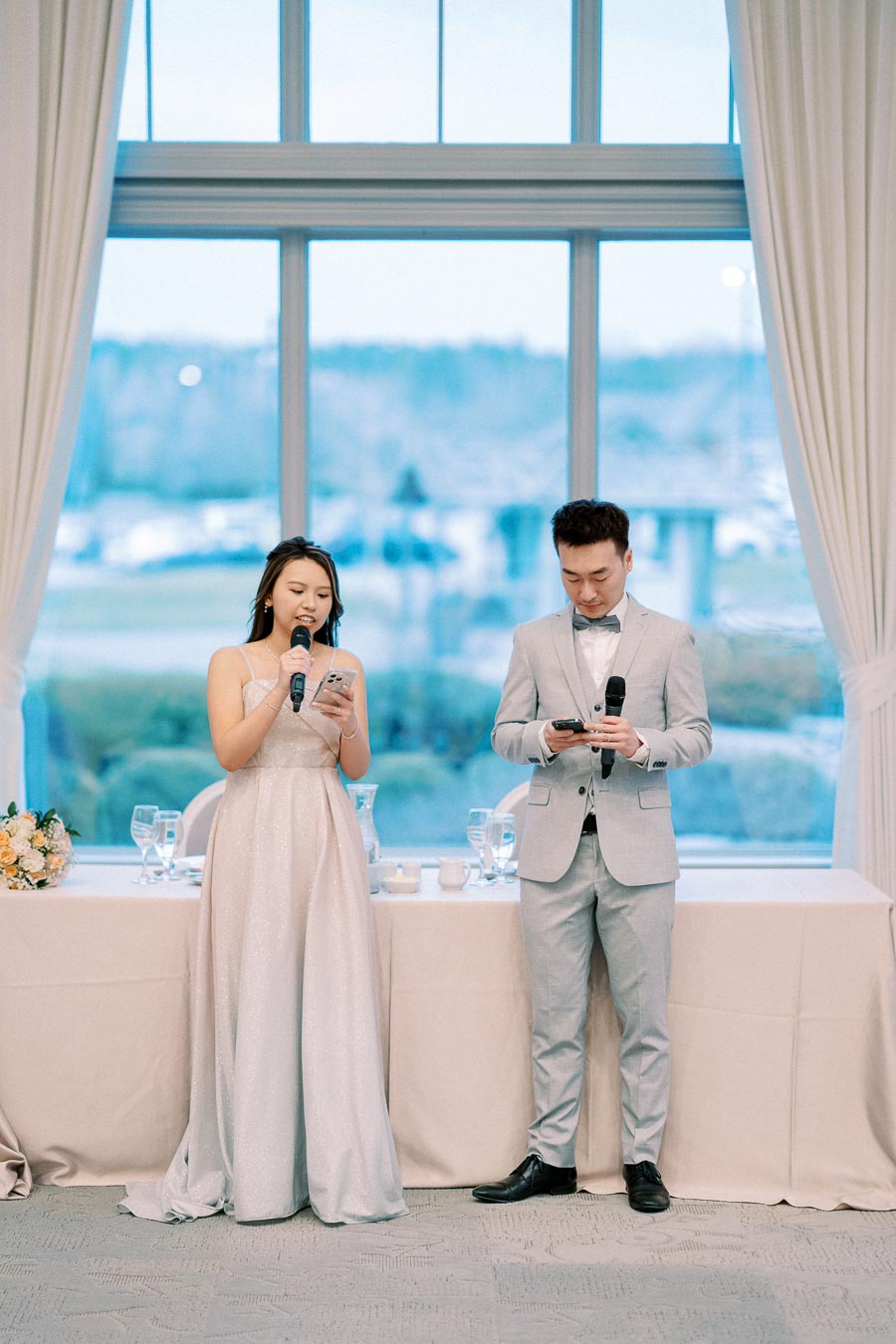 Two elegantly dressed individuals giving a speech at a wedding reception, standing in front of a large window with scenic views.