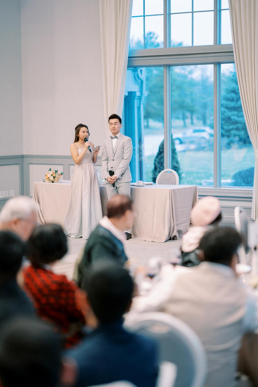 A couple gives a speech at a wedding reception, standing by a table with floral decor, in a bright room with large windows and gathered guests.