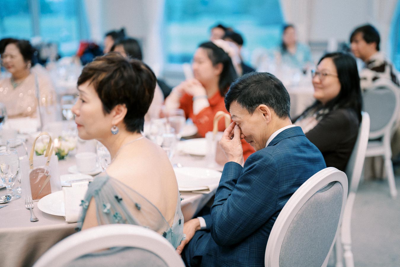 Guests seated at a formal event, with a man in a blue suit rubbing his eyes and other attendees listening attentively, creating an atmosphere of engagement and emotion.