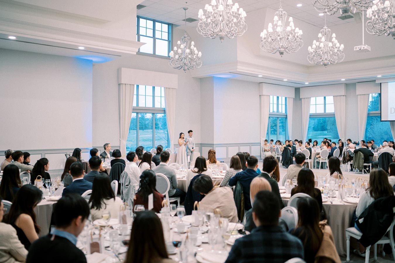Wedding reception in an elegant banquet hall with chandeliers, featuring a speaker addressing seated guests at round tables.