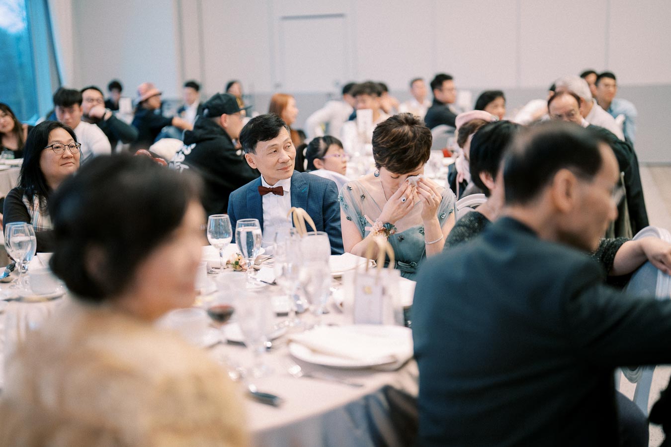 Wedding reception scene with elegantly dressed guests seated at round tables, sharing conversations and experiencing emotional moments, with soft ambient lighting.