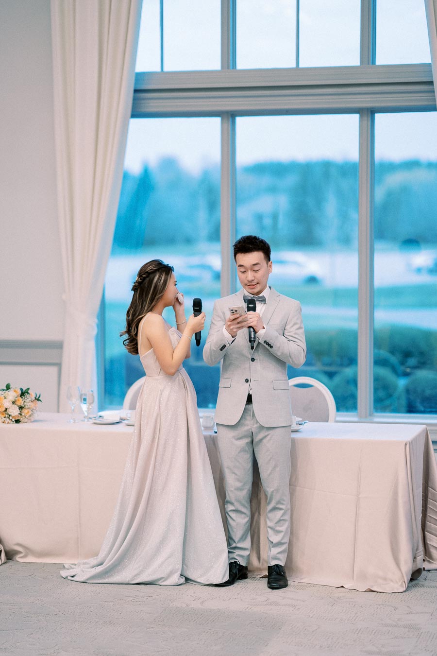 Wedding couple giving heartfelt speeches indoors, woman in elegant gown and man in light gray suit holding microphones near decorated table.
