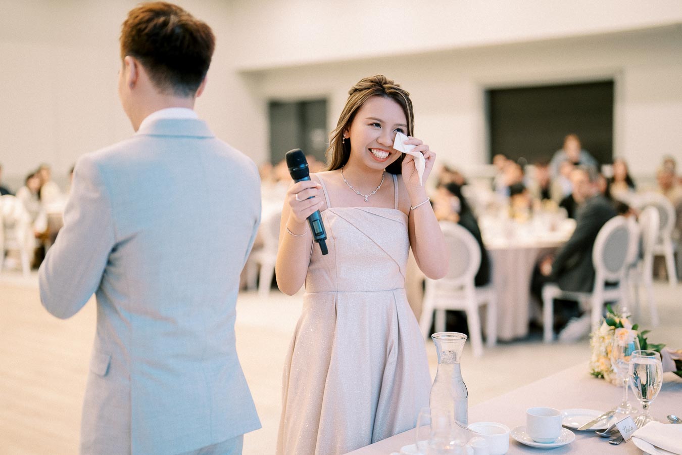 A woman in a formal dress gives a heartfelt speech while holding a microphone and wiping tears at an elegant indoor event.