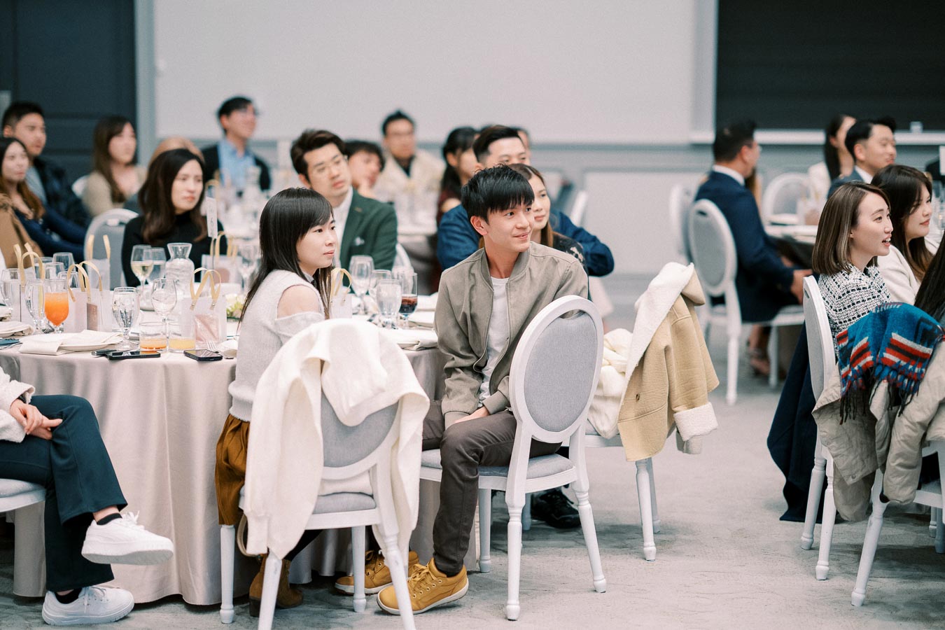 A group of people sitting attentively at round tables during a conference or formal event, with glasses and orange juice on the tables, wearing casual and semi-formal attire.