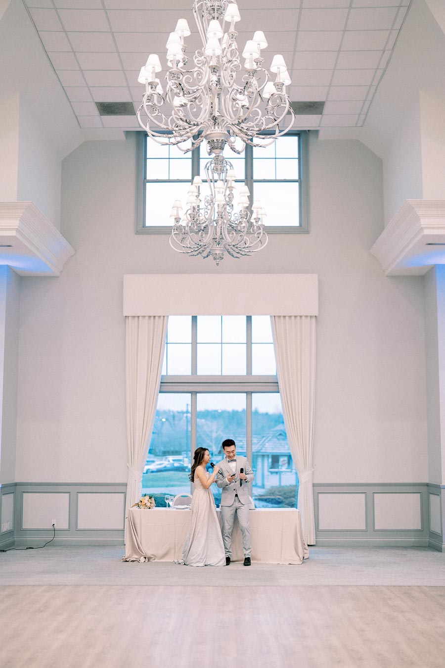 Elegant wedding venue interior with a grand chandelier hanging above a couple standing in front of a large window, dressed in formal attire, sharing a moment during their celebration.