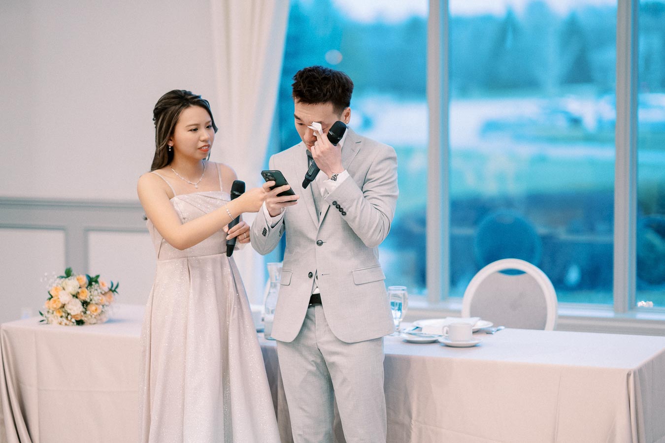A bridesmaid in a shimmering dress hands a microphone to a man in a light gray suit who is wiping tears during a wedding speech, with a flower arrangement and elegantly set reception table in the background.