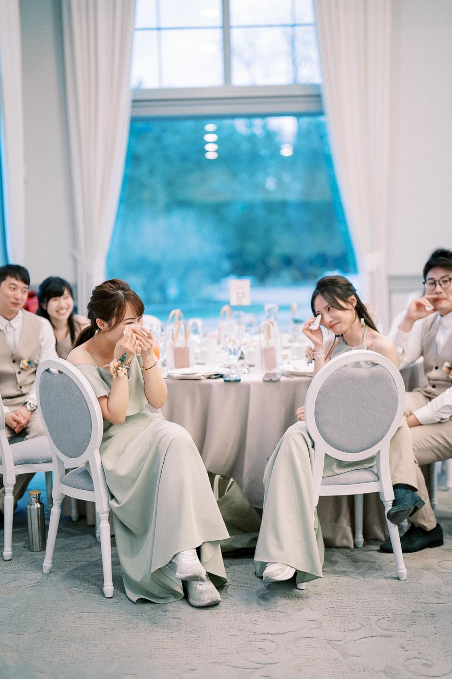 Bridesmaids in matching pastel dresses sitting and laughing at a wedding reception, with elegantly set tables and a large window showcasing a blurred outdoor view in the background.