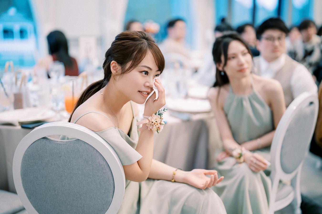 Woman smiling at a wedding reception, dabbing her cheek with a tissue, surrounded by elegantly dressed guests seated at decorated tables.
