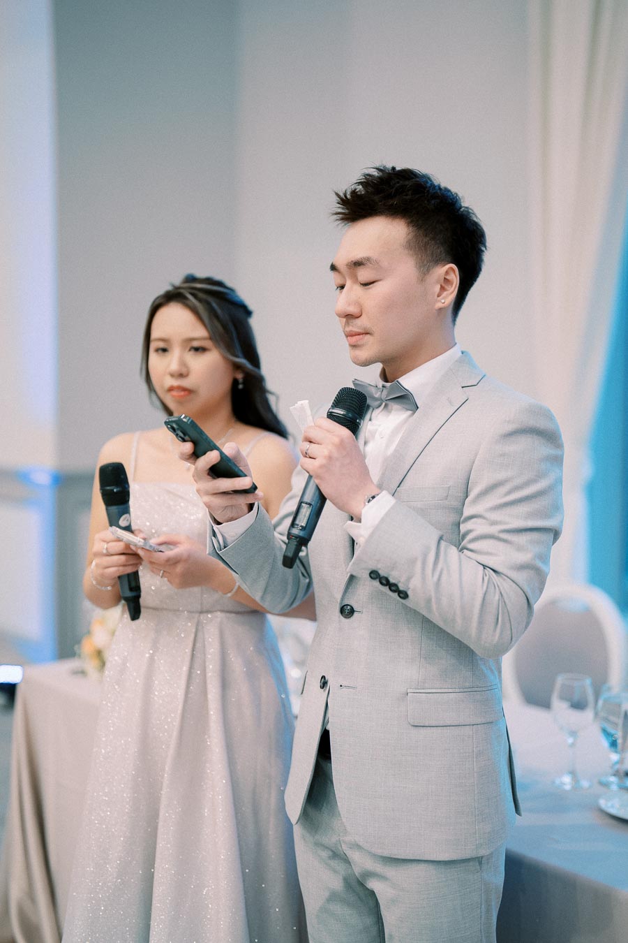 Two individuals in formal attire giving a speech at a wedding reception, holding microphones with a table setting in the background.