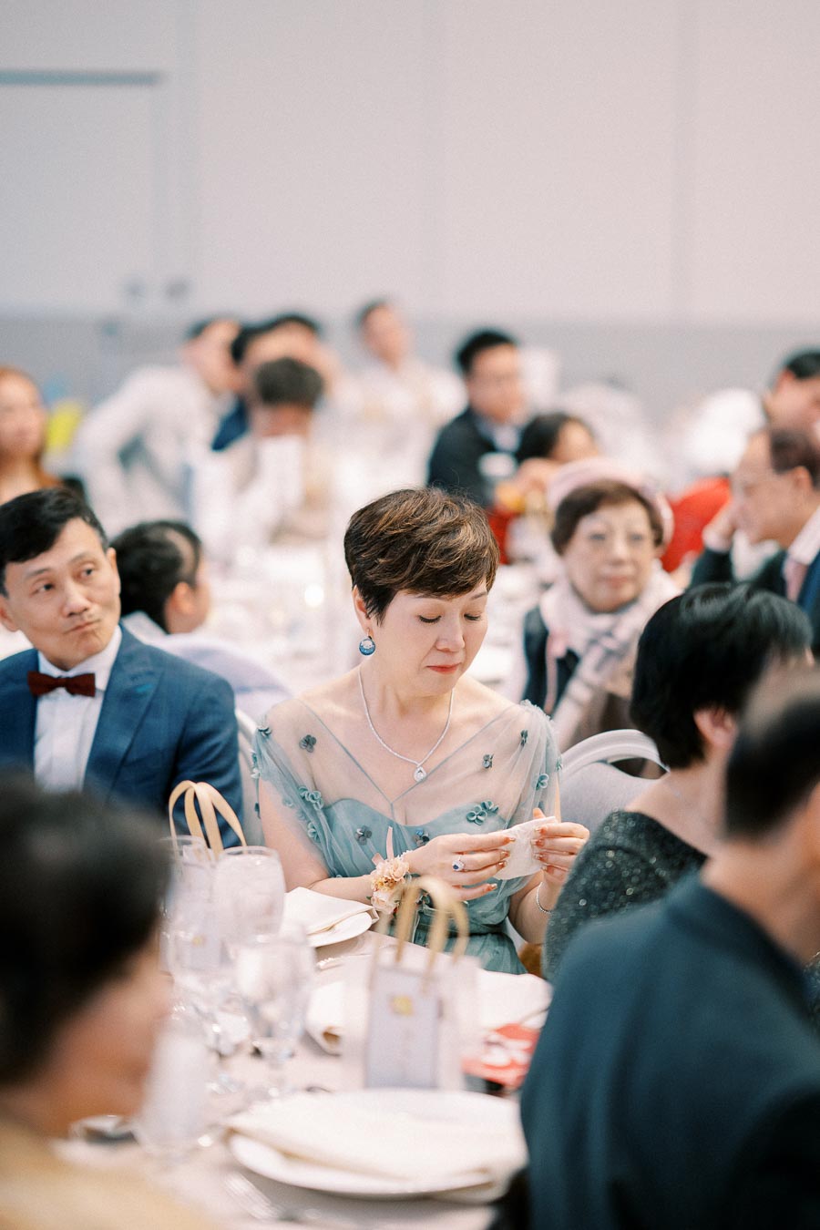 A group of elegantly dressed individuals seated at a formal event, with a woman in a delicate blue dress and a man in a suit and bow tie, surrounded by elegantly set dining tables.