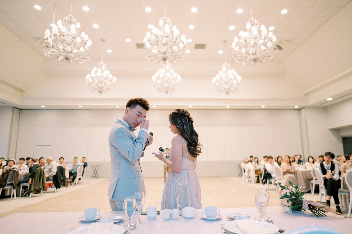 A couple dressed in formal attire sharing emotional vows at a wedding reception, with elegant chandeliers overhead and a seated audience in the background.