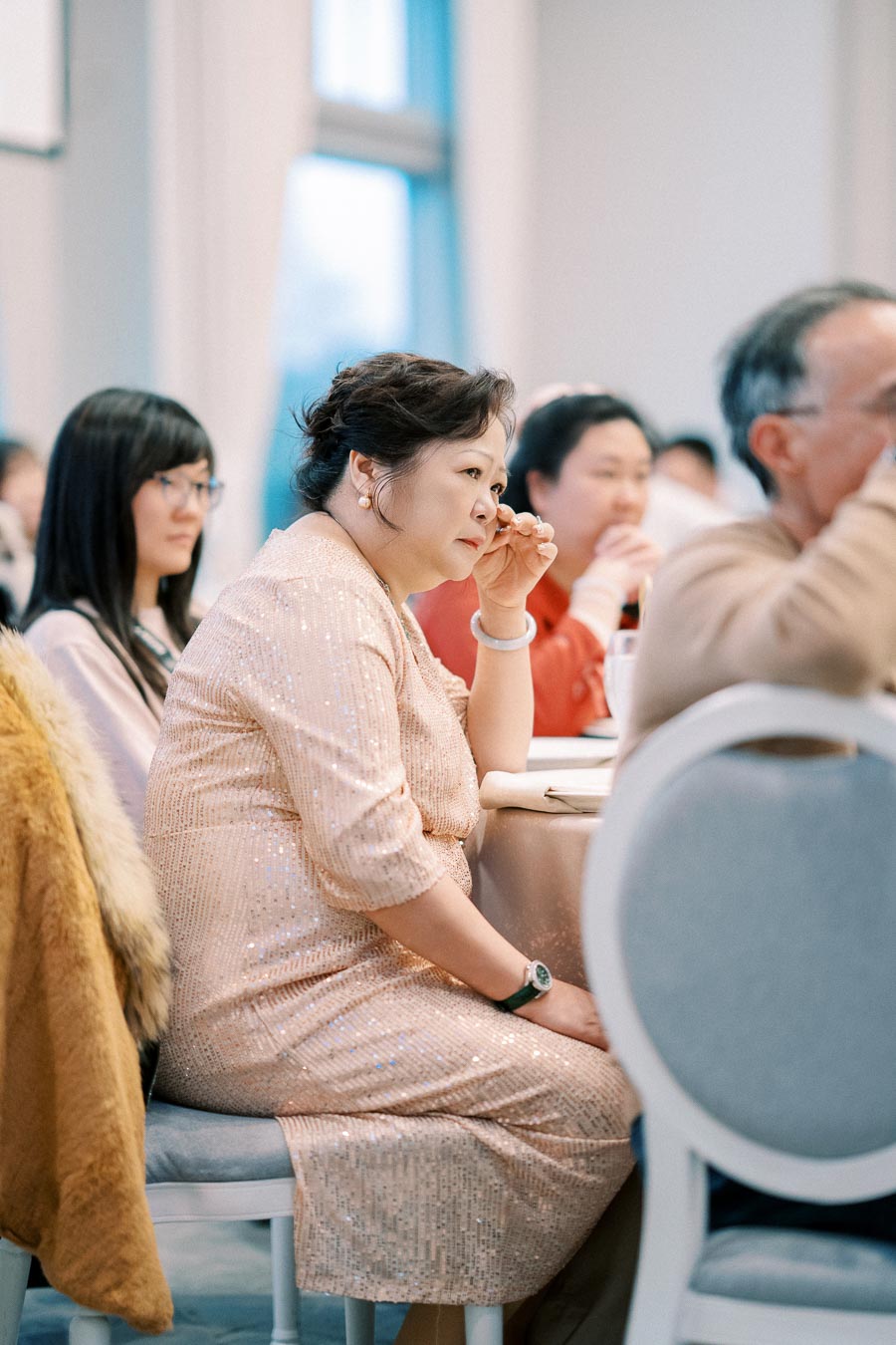 Elderly woman in a sparkly dress attentively listening at an event, seated among a group of people.