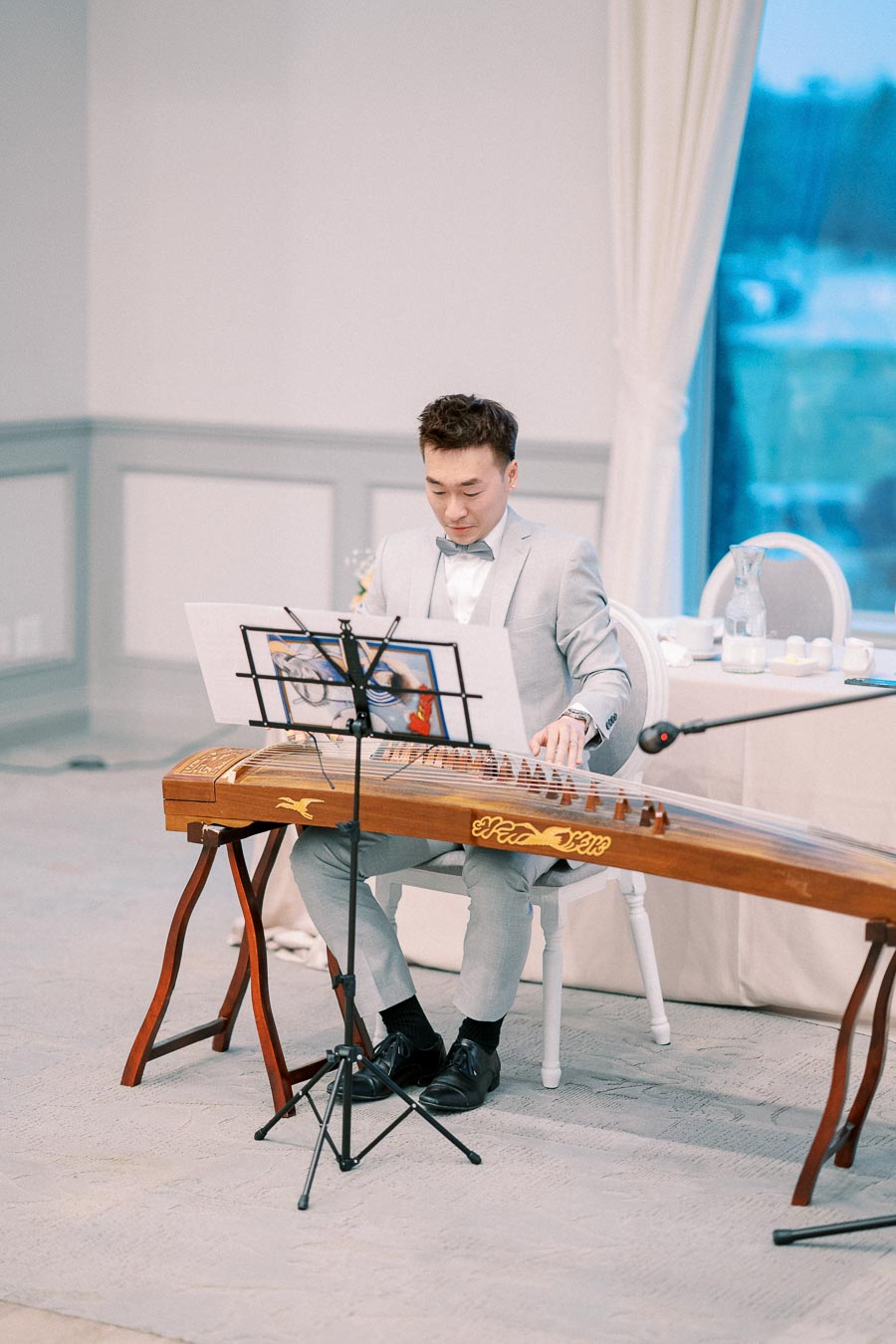 A man in a light grey suit playing a traditional guzheng instrument in an elegant indoor setting, with a music stand in front of him.