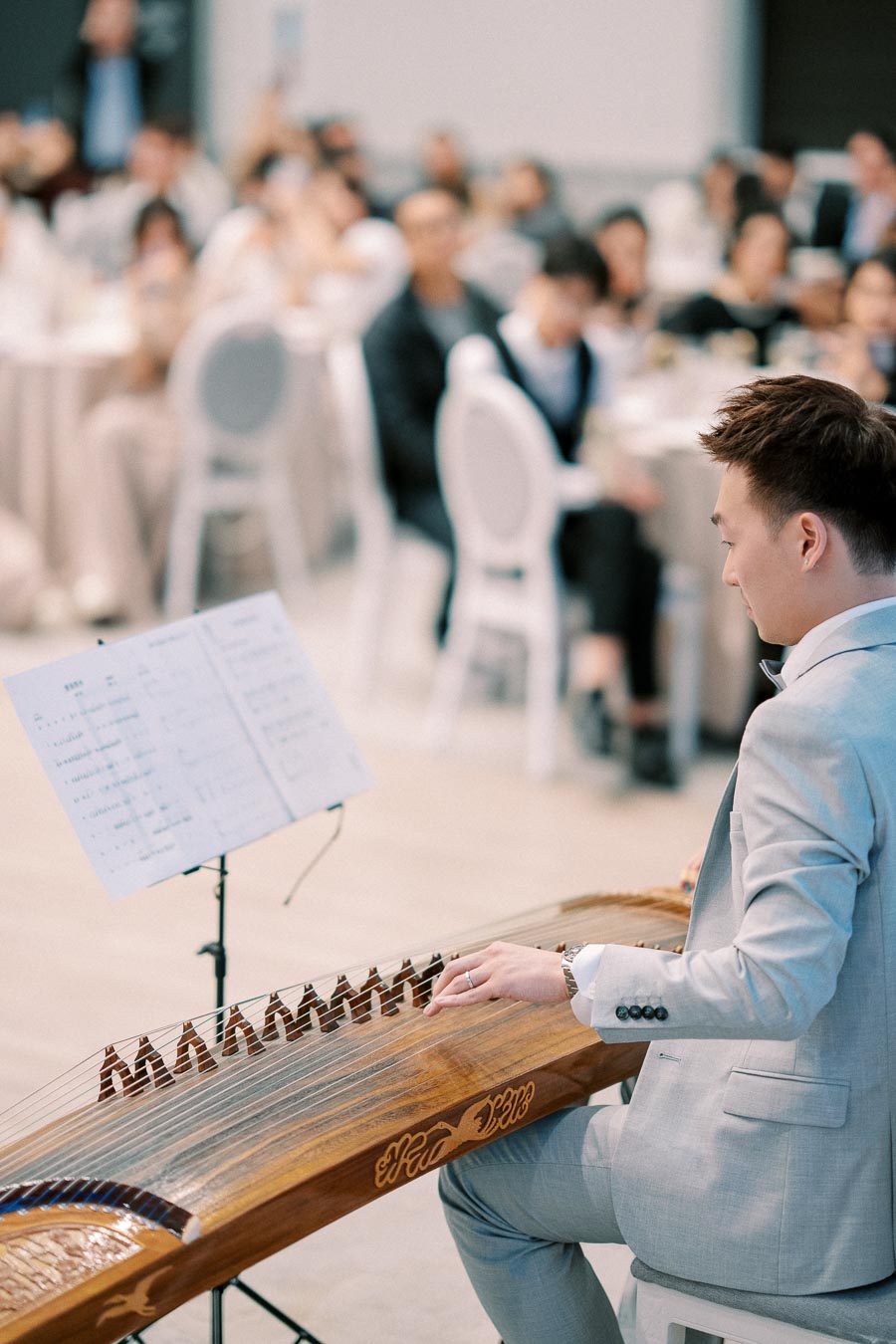 A man in a light gray suit playing a traditional stringed instrument at an indoor event, with an audience in the background, and sheet music on a stand nearby.