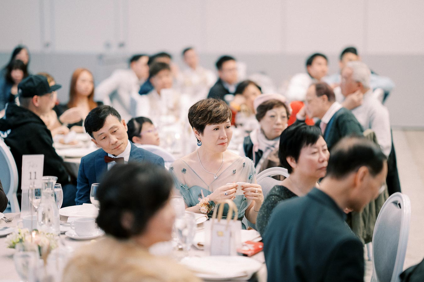A group of elegantly dressed people seated at a formal banquet, attentively listening during the event. Tables are set with white tablecloths, place settings, and floral centerpieces.
