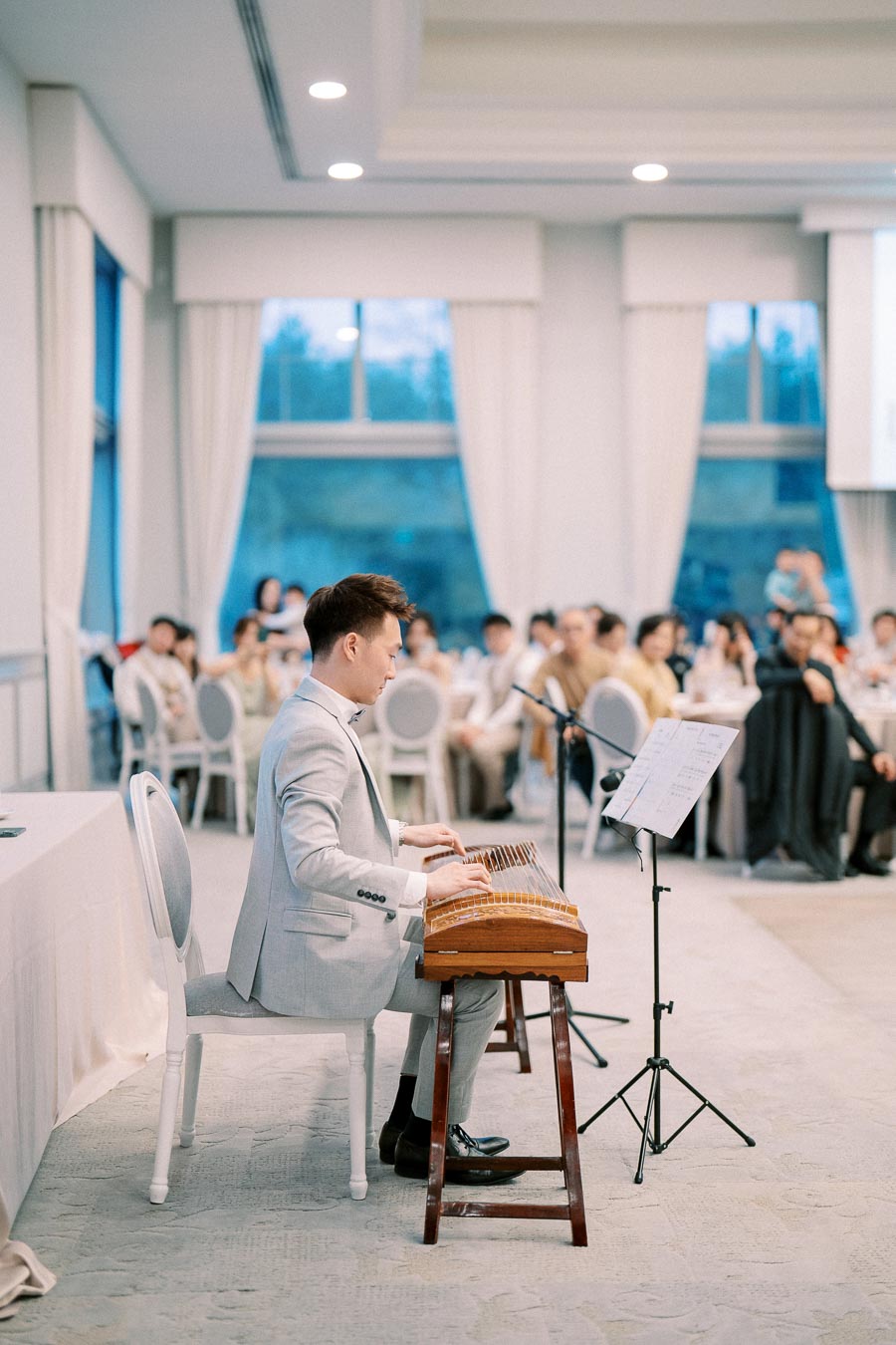 Musician in a grey suit performing on a traditional stringed instrument, with a seated audience inside a bright, elegantly decorated room.