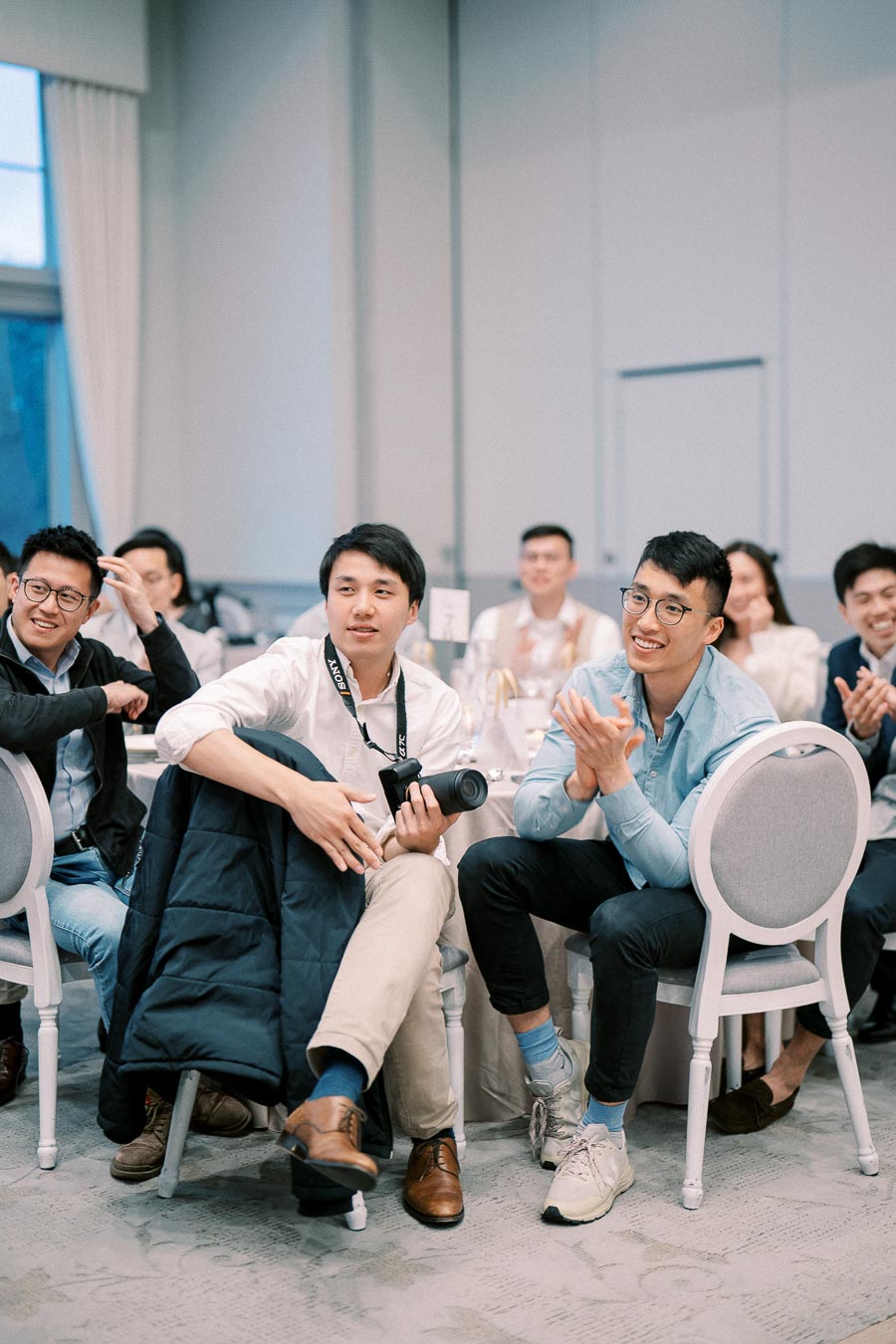 Group of young adults at a formal event, seated around a table, attentively watching a presentation. One person holding a camera, others smiling and clapping, dressed in casual business attire.