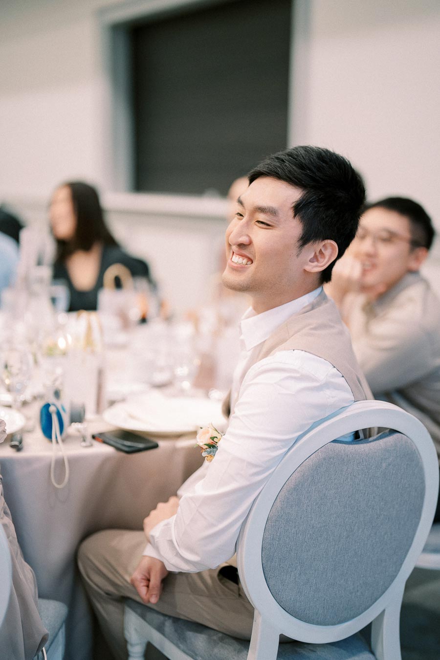 Man smiling at a formal event, seated at a decorated table with others in the background, creating a joyful atmosphere.