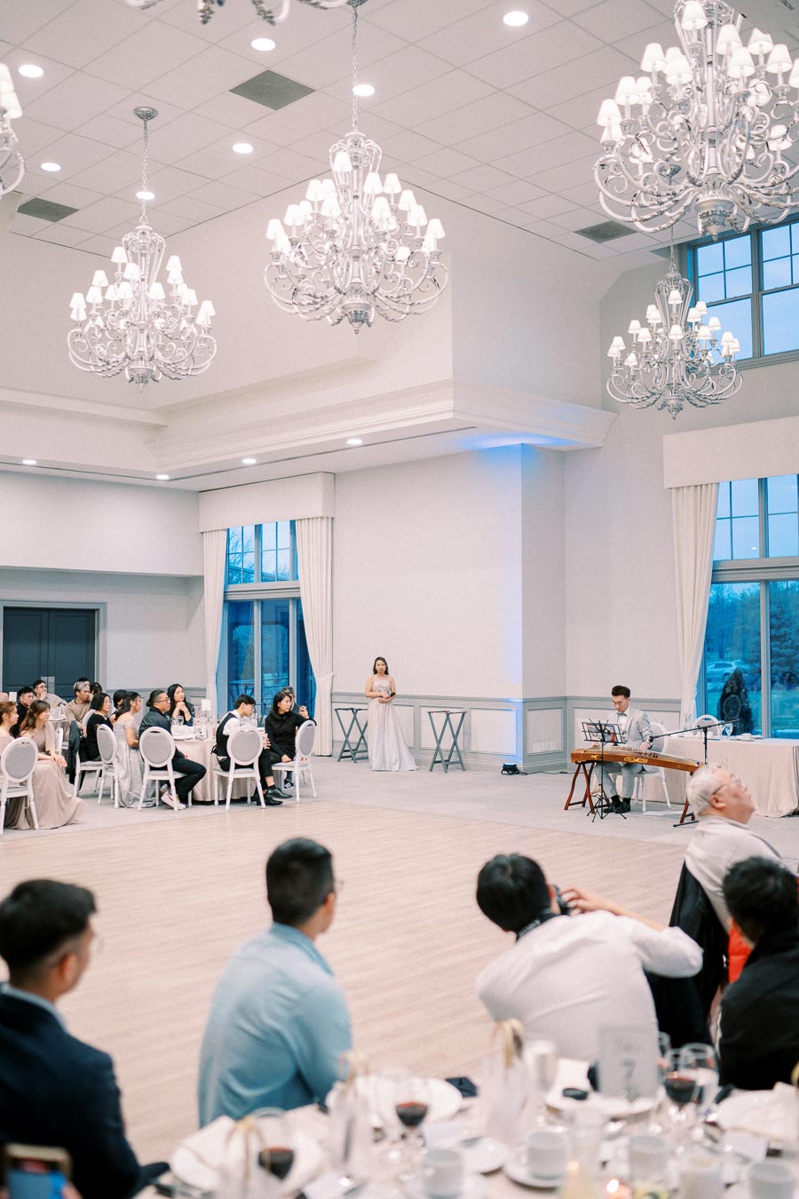 Elegant banquet hall with chandeliers, guests seated at tables, a musician playing an instrument, and a woman in a gown speaking at a formal event.