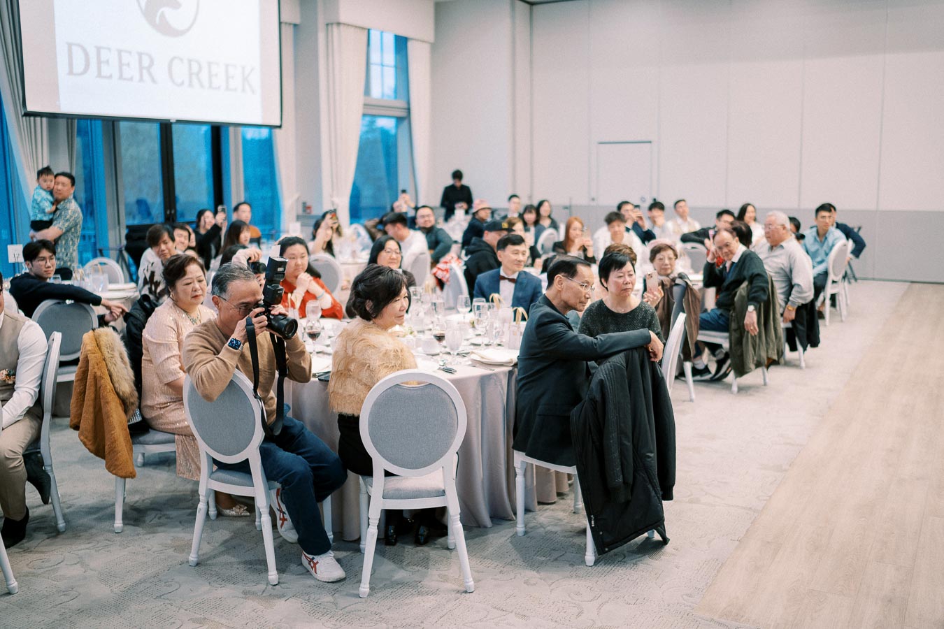 A large group of people seated at round tables during a formal event at Deer Creek event venue, with a photographer capturing the moment in the foreground.