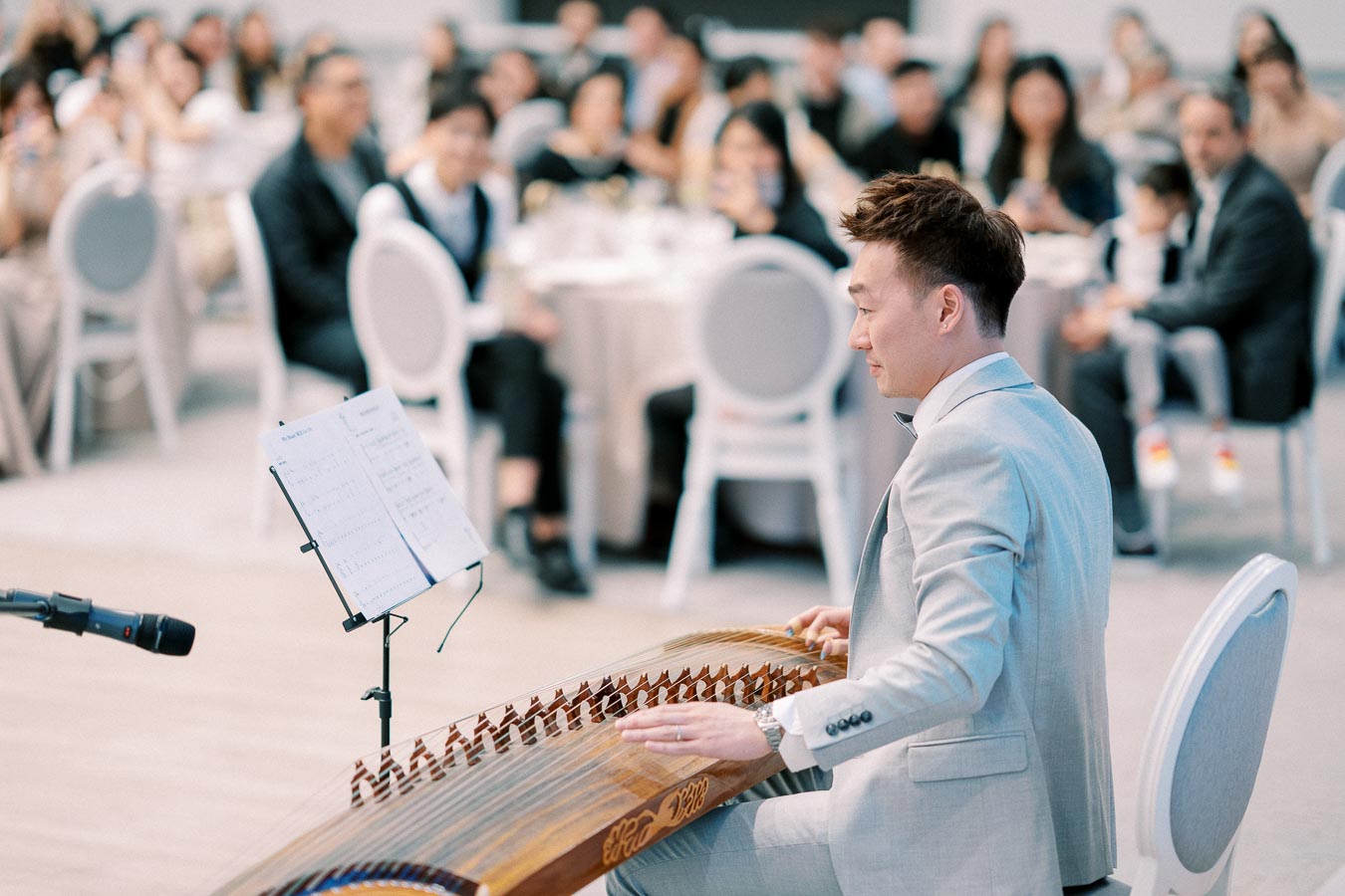 A musician in a light gray suit playing a traditional string instrument at an event, with an audience attentively watching in the background.
