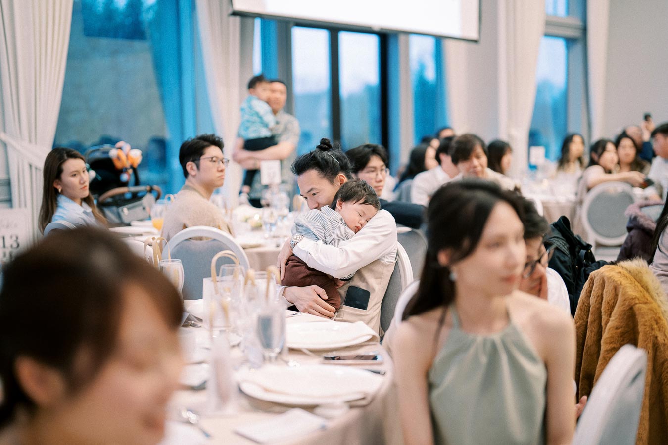 A warm family gathering with adults and children seated around elegantly set dining tables, featuring a father gently cradling his sleeping child while surrounded by attentive guests.