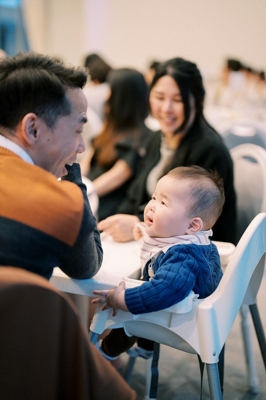 Baby sitting in a high chair interacting with a smiling adult in a cozy social gathering setting.