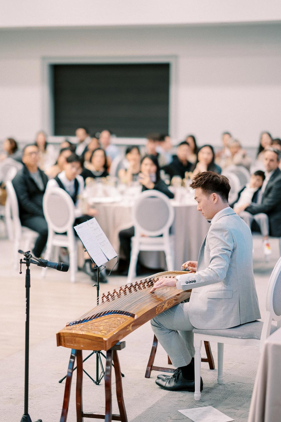 Man in a light suit playing a traditional string instrument at a formal event, with an audience seated at tables in the background.
