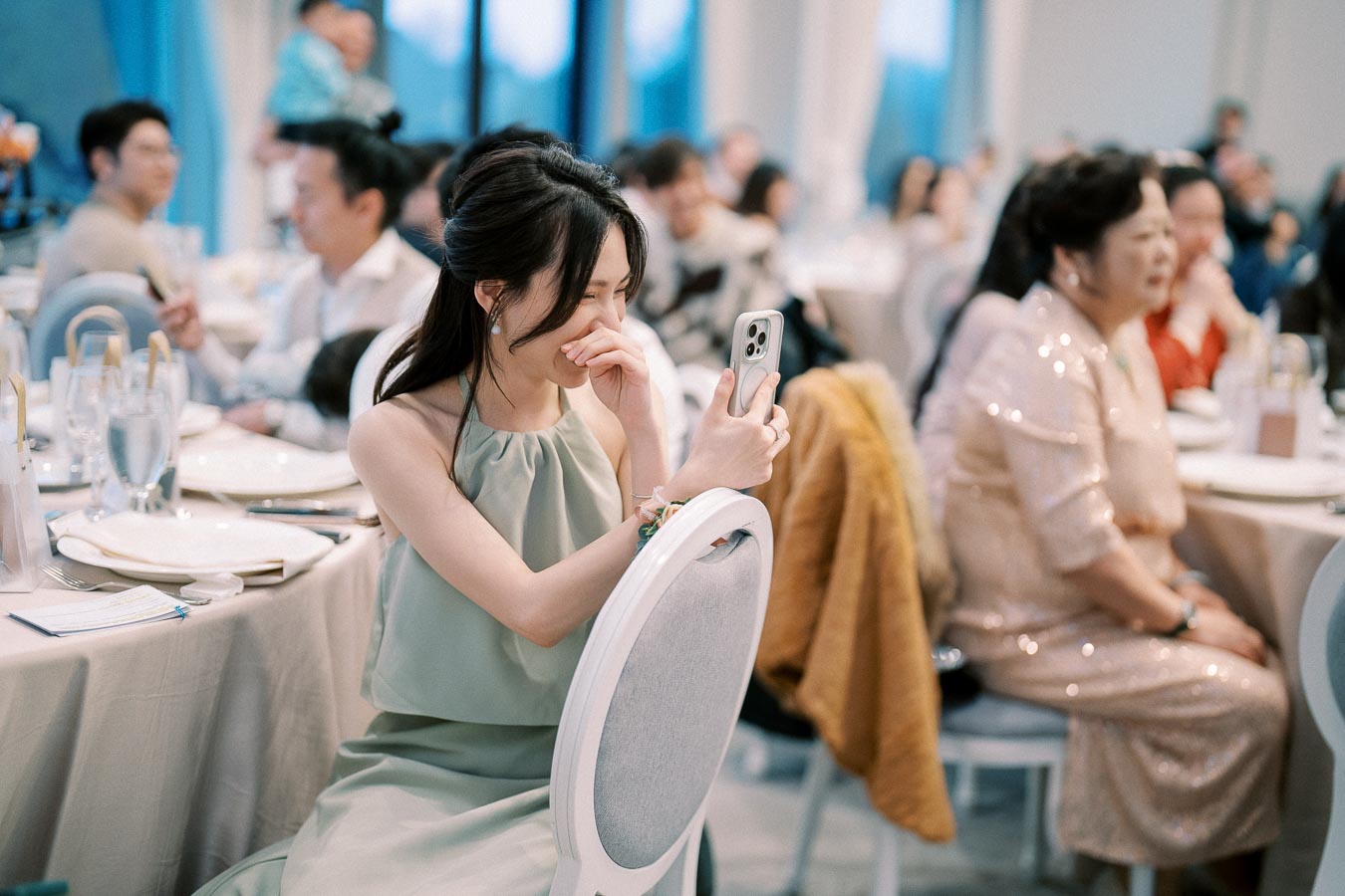 Young woman in a green dress smiling and taking a selfie at an elegant indoor event. The room is filled with seated guests in formal attire, with decorated tables in the background.