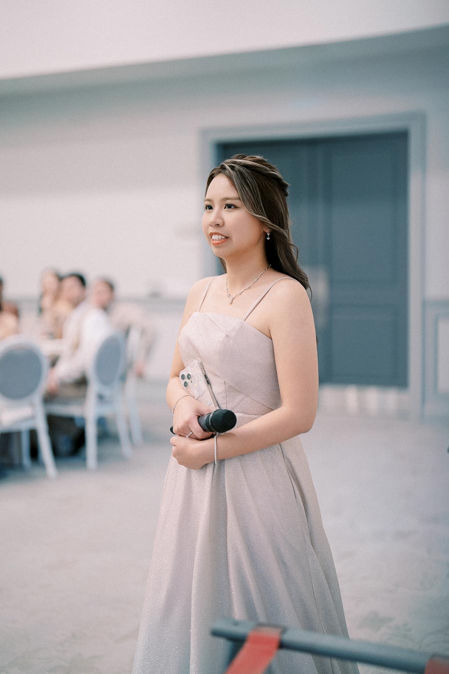 A woman in an elegant strapless dress holds a microphone and phone, standing in an indoor setting with blurred guests seated in the background.