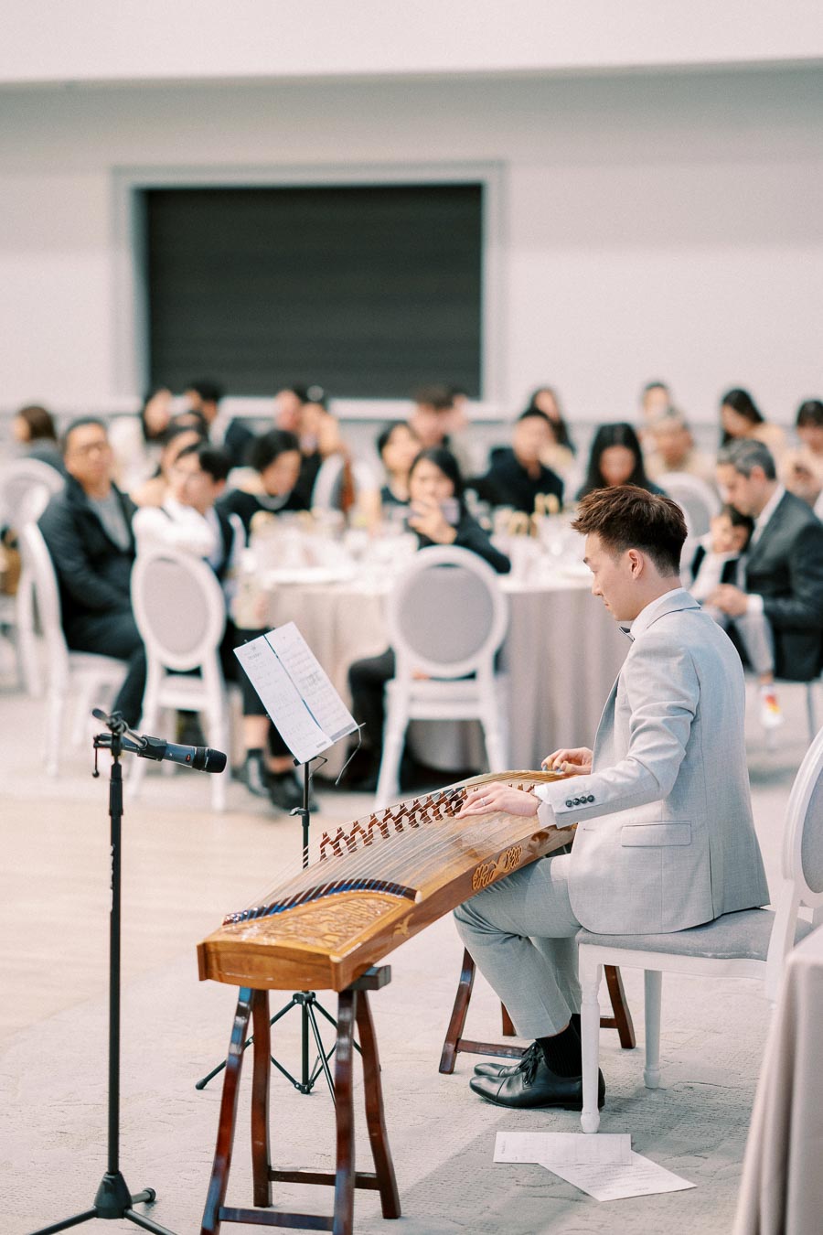 A musician in a gray suit plays a traditional stringed instrument at a formal event, with an attentive audience seated at round tables in the background.
